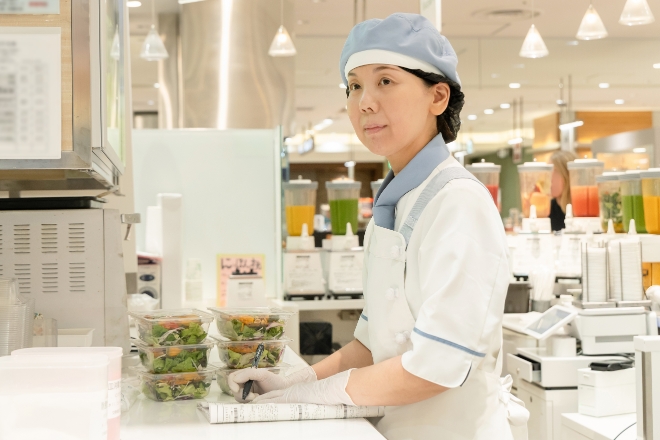 惣菜売り場のカウンターで、サラダについて記録を取るスタッフ