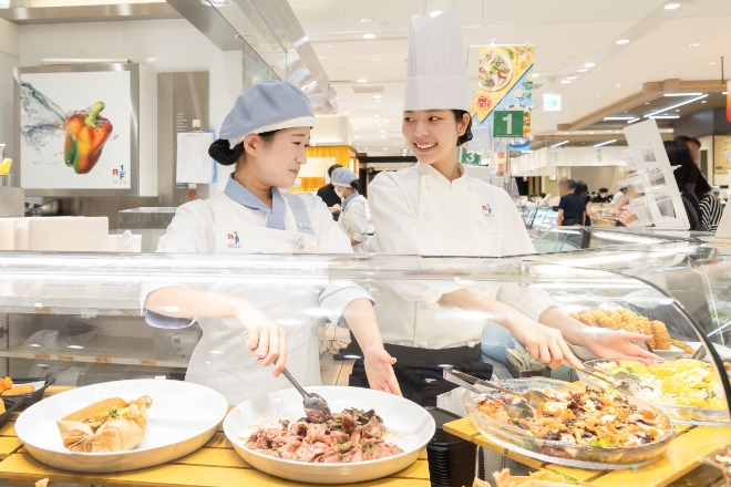 惣菜売り場のショーケース前で、料理を確認しながら会話するスタッフ2名
